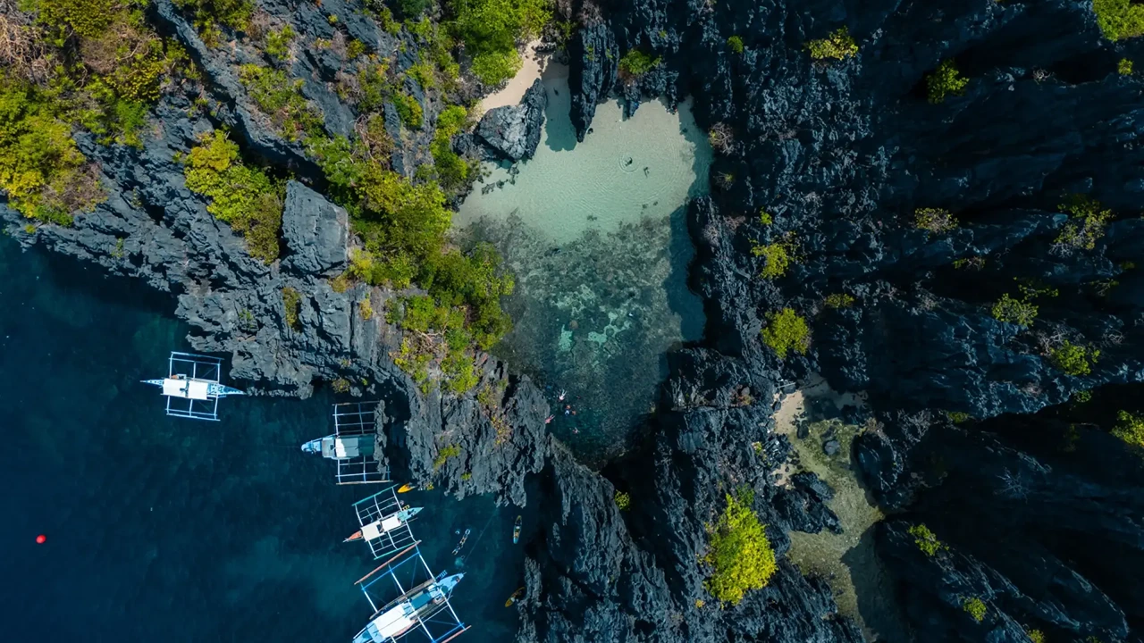 Top-down aerial of a hidden pool enclosed by jagged karst with outrigger boats at its edge, El Nido