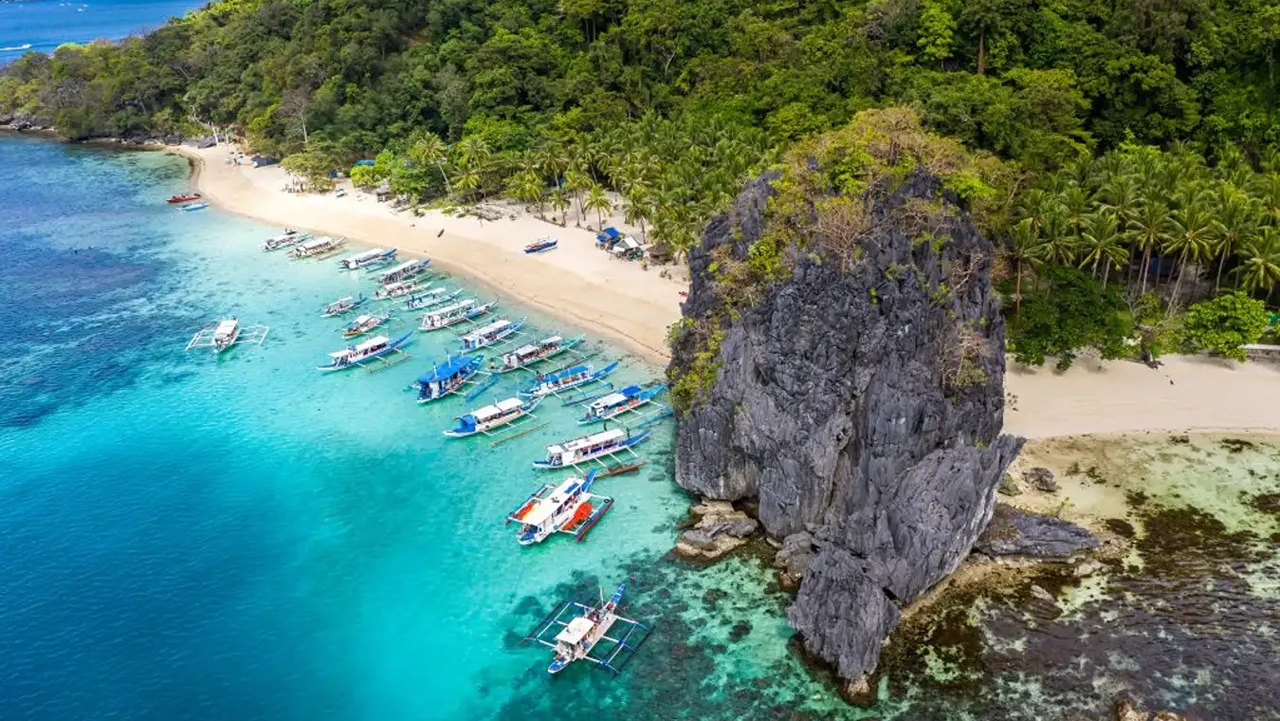 Aerial view of a beach with a massive karst monolith and a row of moored outrigger boats, El Nido