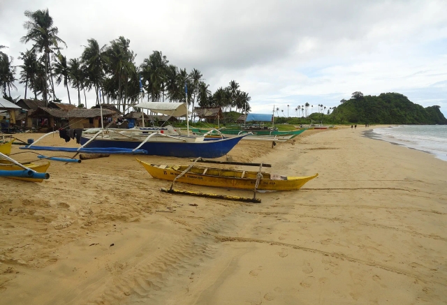 Outrigger boats and nipa huts on the wide sand of Nacpan Beach, El Nido, under an overcast sky