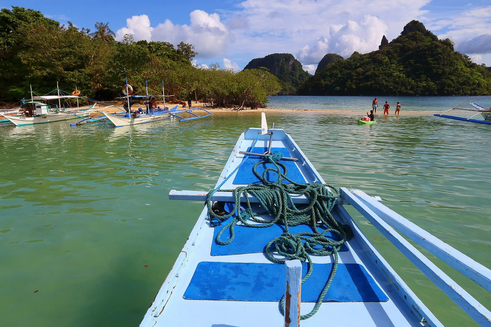 Bow of an outrigger boat with coiled rope approaching a sandbar with moored boats near El Nido