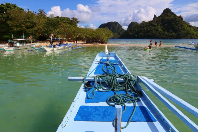 Bow of an outrigger boat with coiled rope approaching a sandbar with moored boats near El Nido