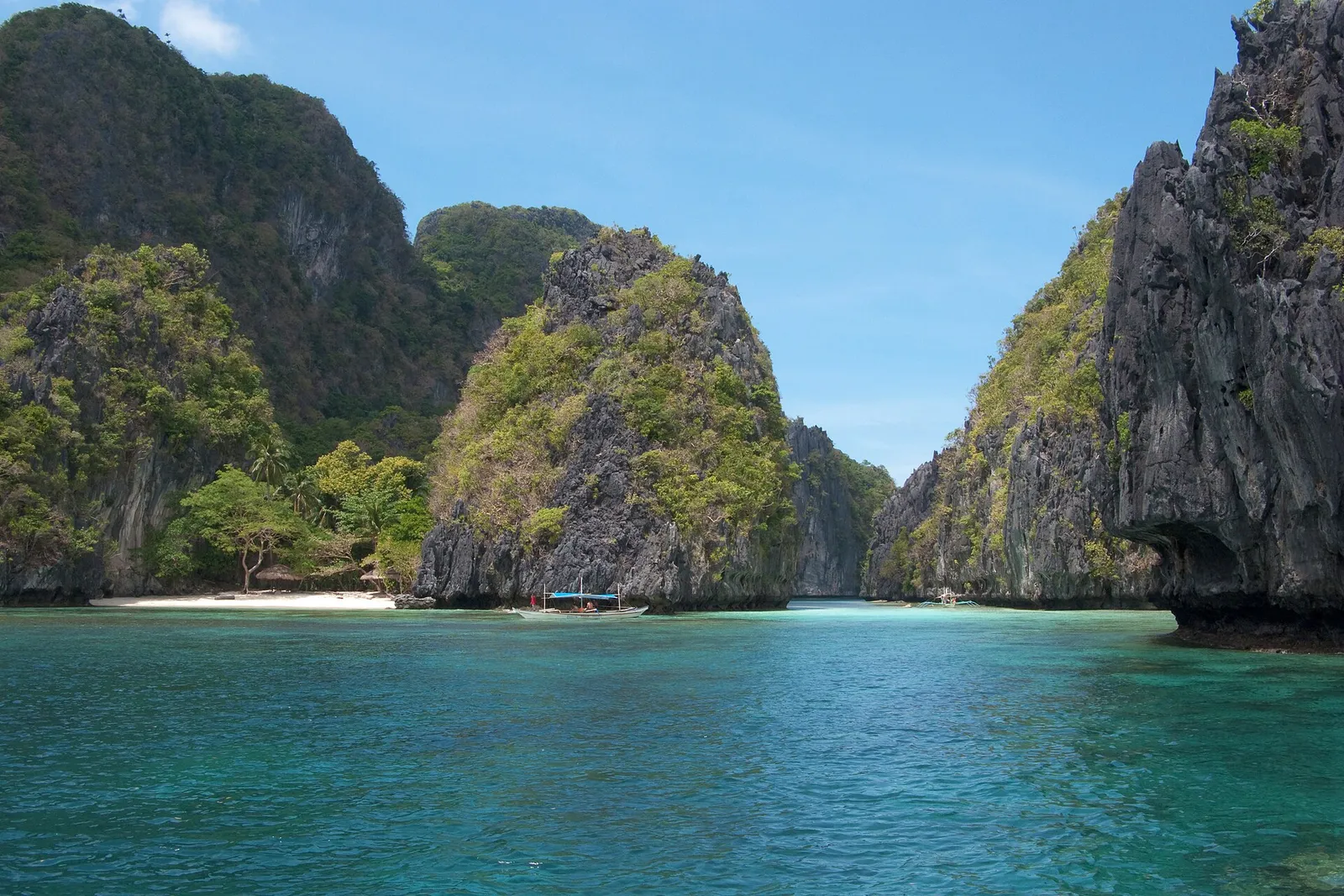 Karst islands and a small beach with huts and an outrigger boat on calm water in Bacuit Bay, El Nido