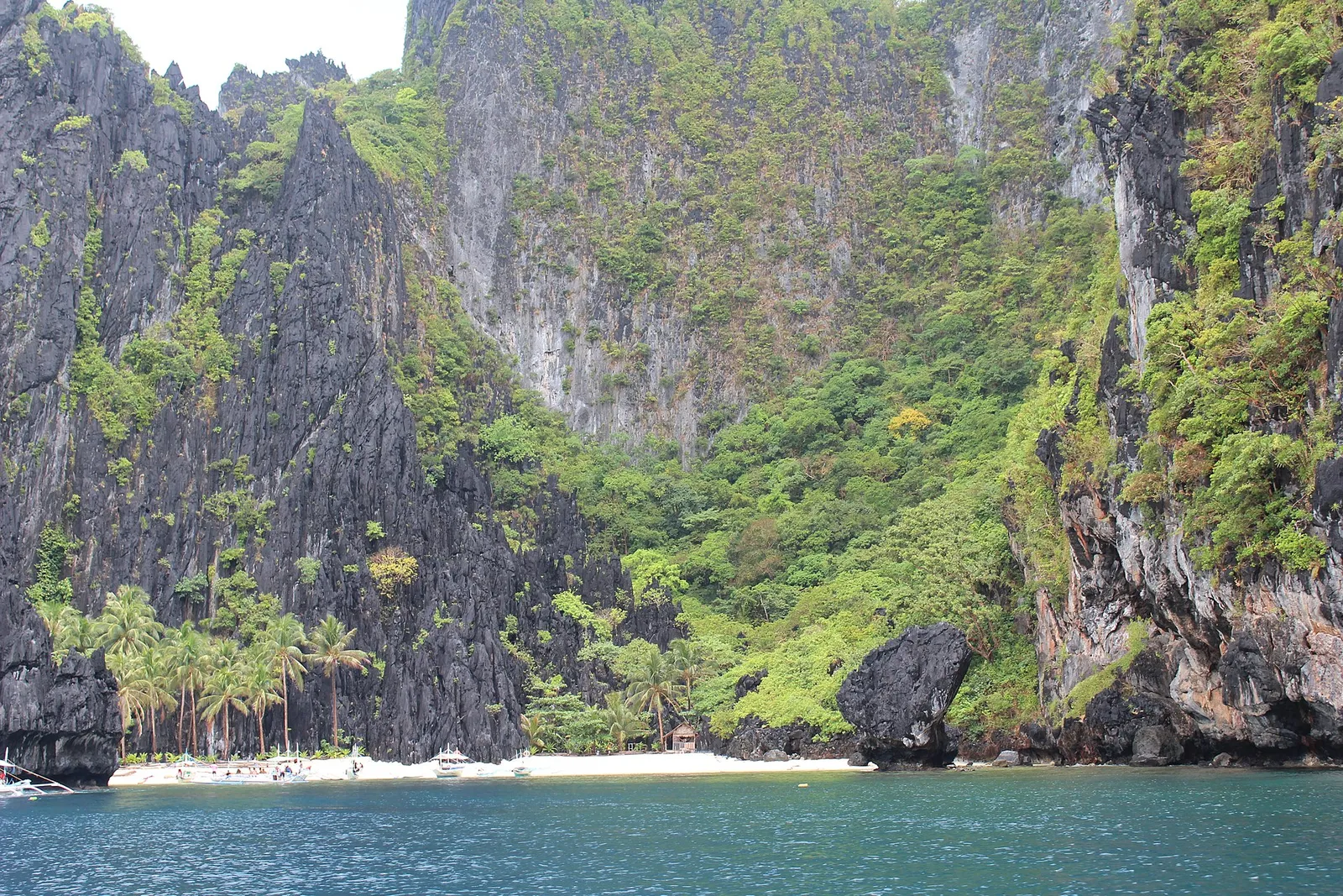 Small white beach with palms and outrigger boats at the foot of a towering karst wall near Secret Beach, El Nido