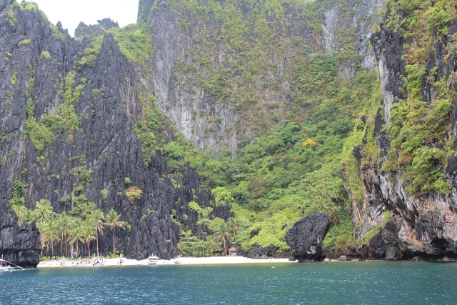 Small white beach with palms and outrigger boats at the foot of a towering karst wall near Secret Beach, El Nido