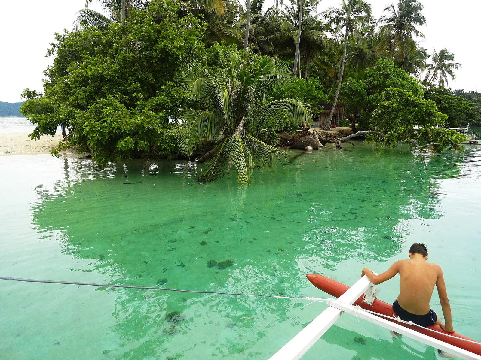 Boy sitting on the red outrigger float of a bangka over clear green shallows near German Island, Port Barton