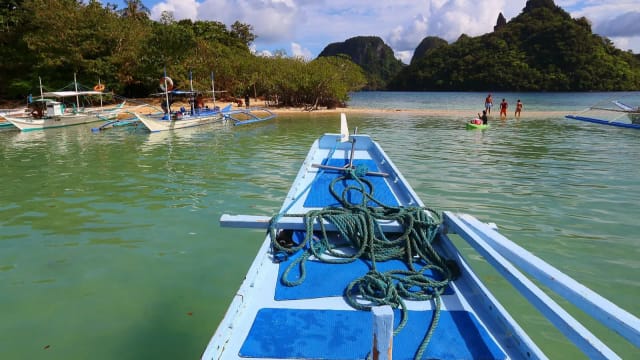 Bow of an outrigger boat with coiled rope approaching a sandbar with moored boats near El Nido
