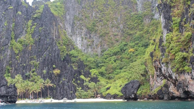 Small white beach with palms and outrigger boats at the foot of a towering karst wall near Secret Beach, El Nido