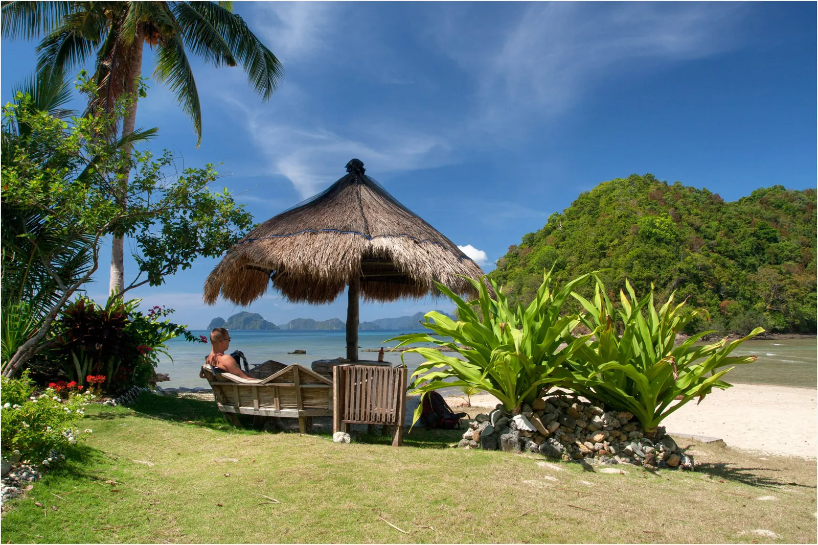 Traveller resting under a thatched beach umbrella at Las Cabanas Beach, El Nido, with Bacuit Bay islands on the horizon