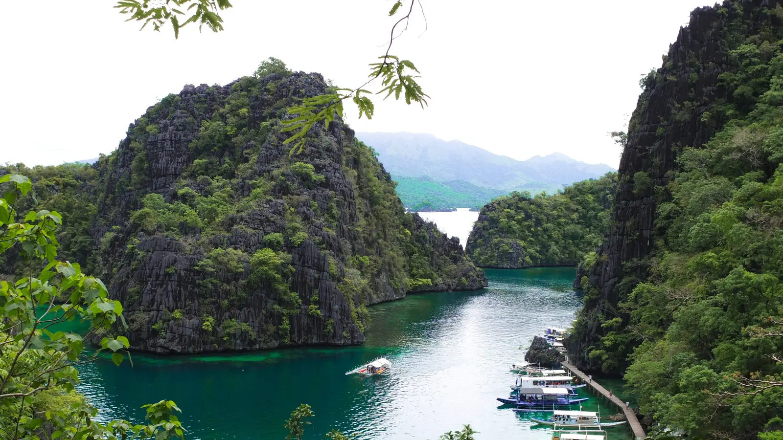 Jagged limestone karst rising over the green water of the Kayangan Lake cove, with a boardwalk and moored boats, Coron Island