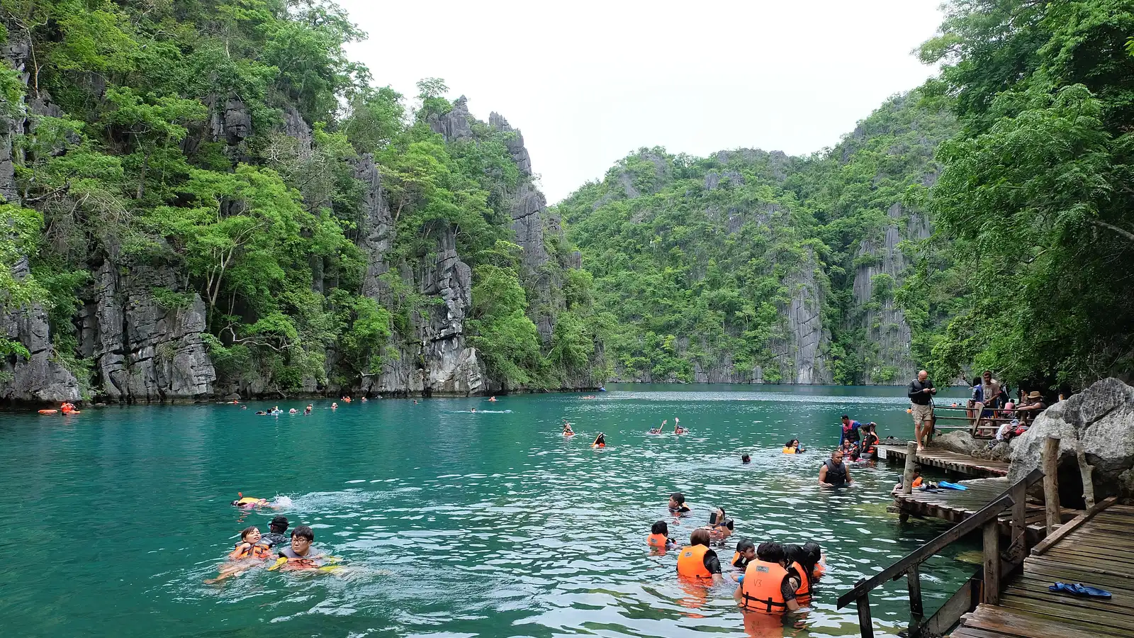 Swimmers in life vests in the clear turquoise water of Kayangan Lake beneath forested limestone cliffs, Coron
