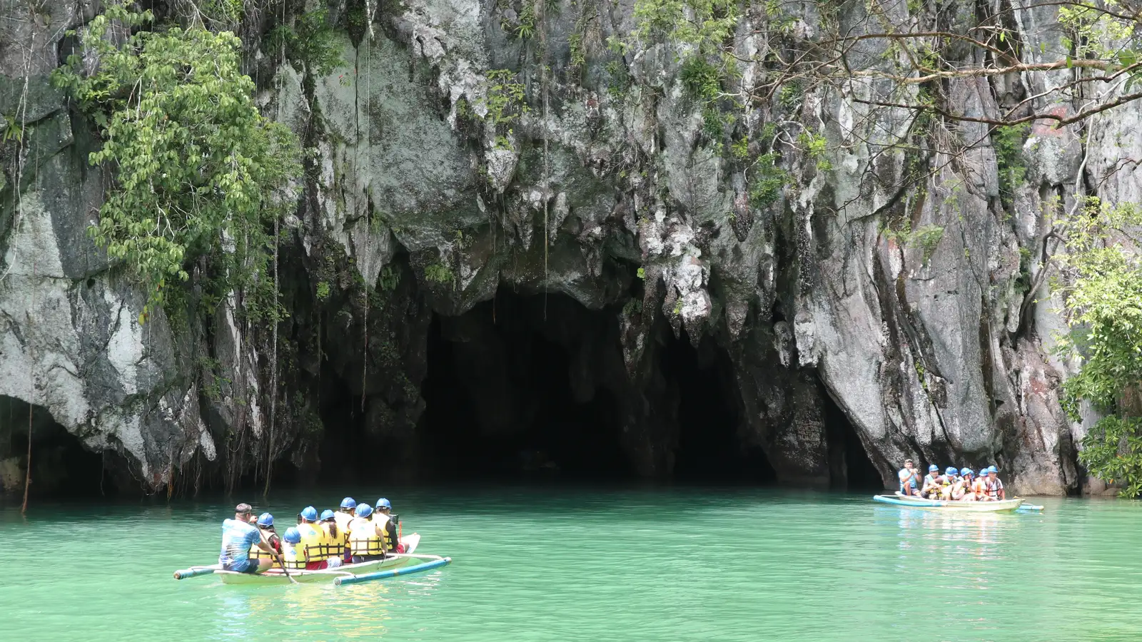 Tour boats with helmeted visitors in life vests paddling toward the Underground River cave mouth beneath a sheer limestone cliff, Sabang